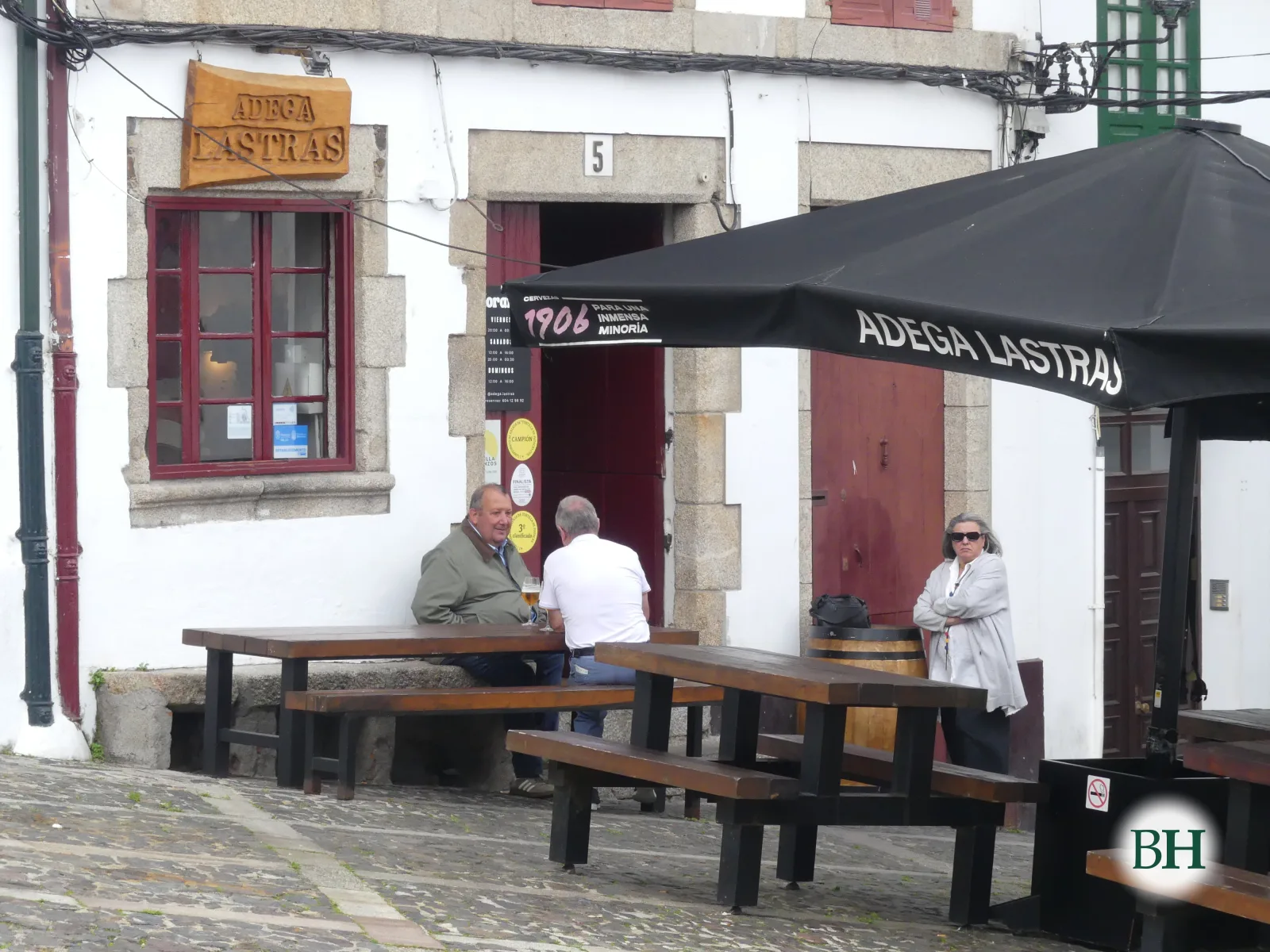 Fachada branca da Adega Lastras, co letreiro de madeira, xanela e porta vermellas, e mesas de madeira na rúa baixo un parasol negro co nome do local. Dous homes sentados e unha muller de pé.