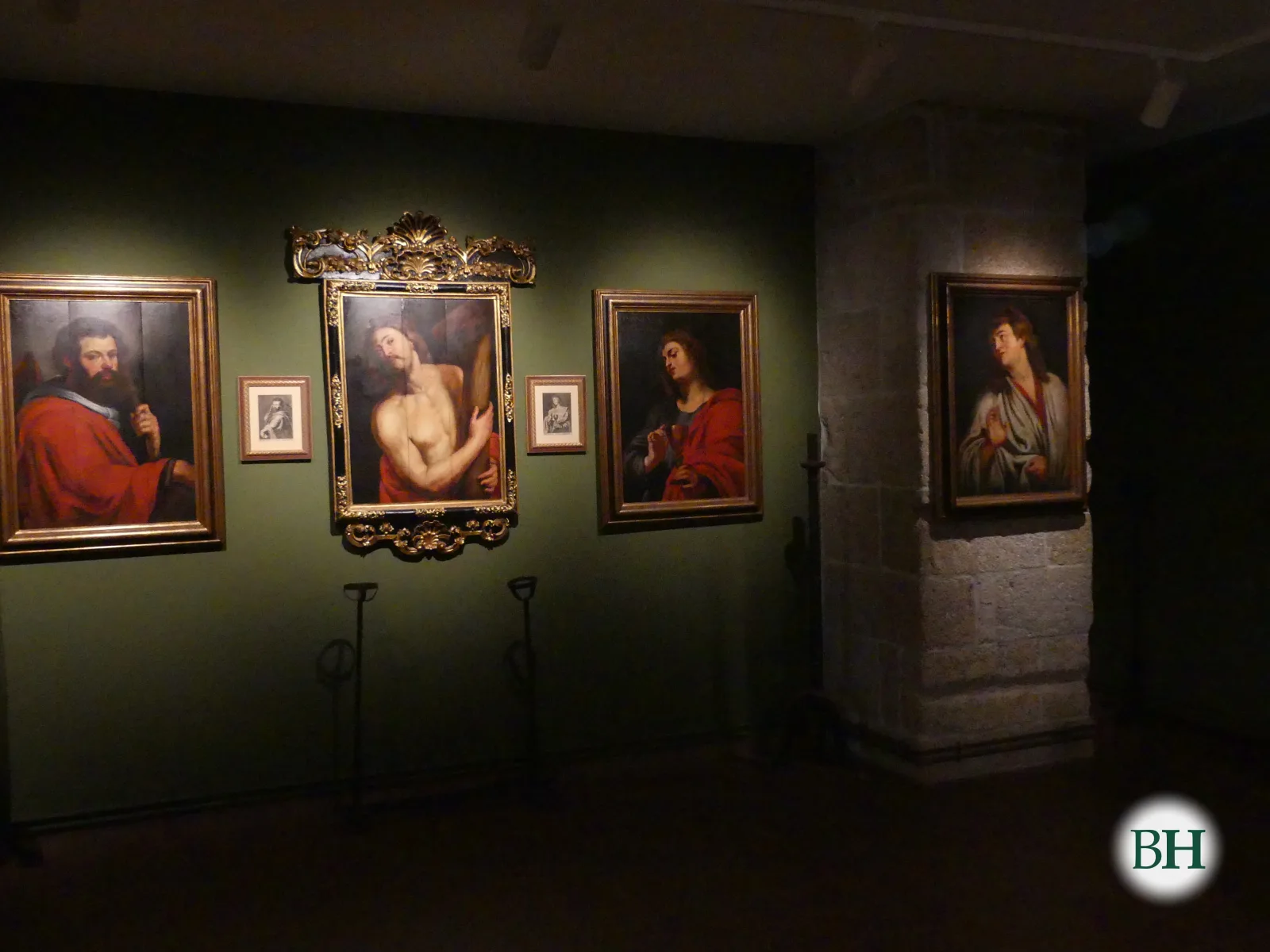 Sala de paredes verdes e pouca luz no Museo das Mariñas con catro óleos de apóstolos e un Cristo coa cruz en marcos dourados, xunto a un piar de pedra.
