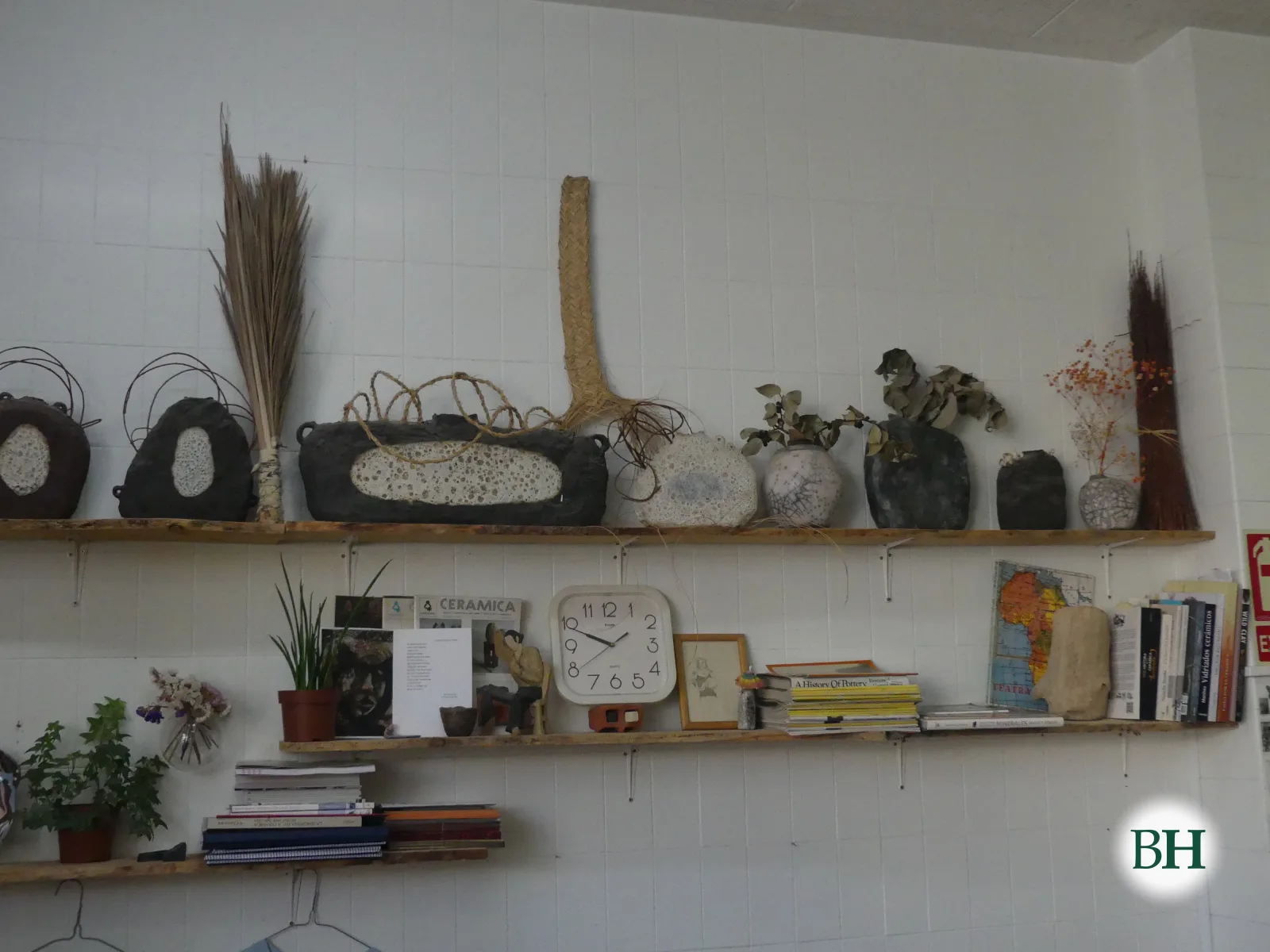 Andel cunha fila de pezas de cerámica escuras e brancas con vimbio, esparto e ramas secas enriba, sobre outro andel con libros, plantas e un reloxo.