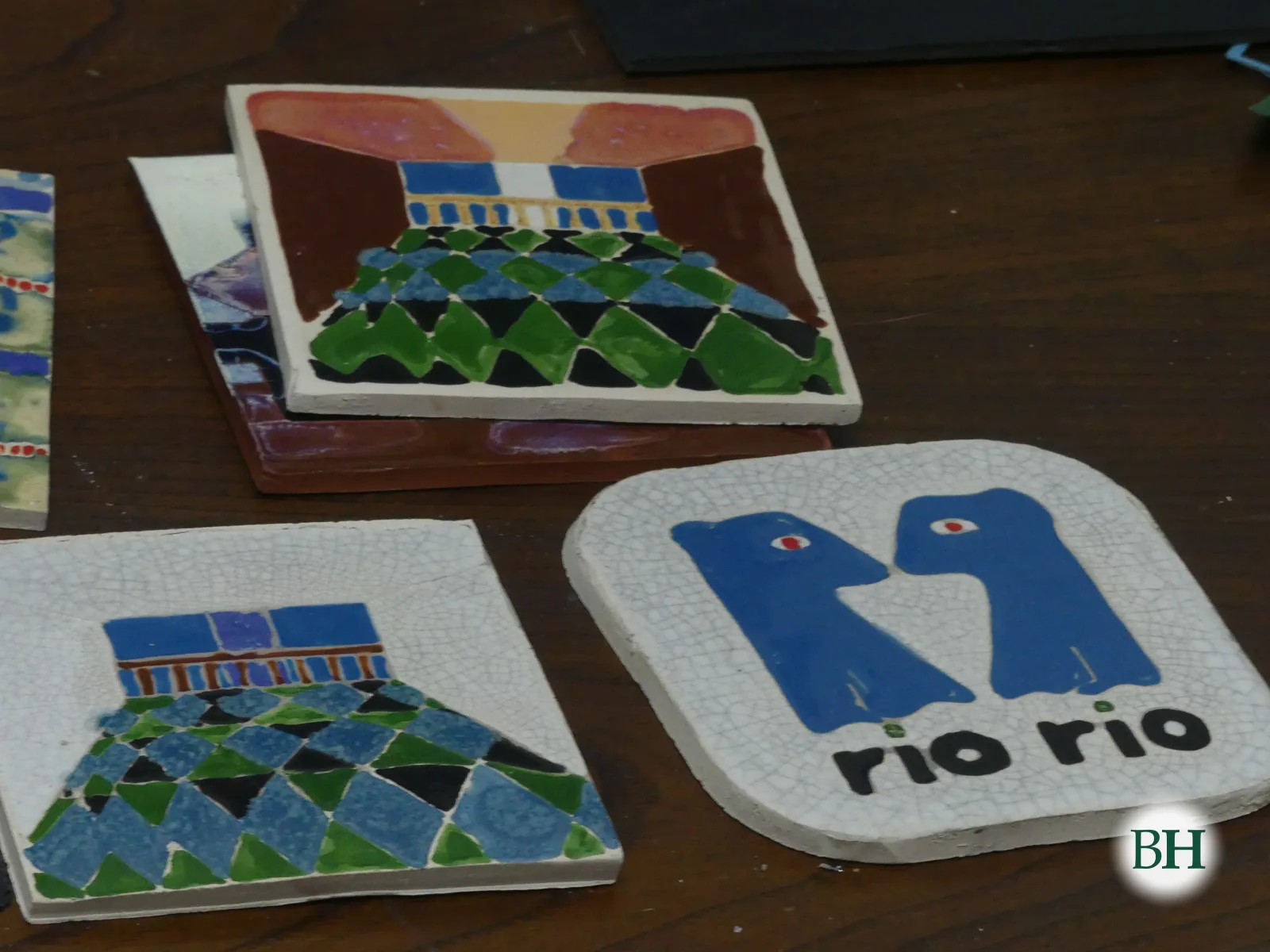 Catro azulexos esmaltados sobre unha mesa de madeira: dous con rombos verdes, azuis e negros baixo un balcón azul, e un cadrado branco co logotipo azul de Río Río, dúas figuras enfrontadas.