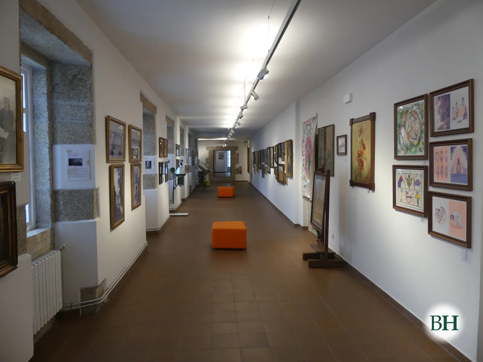 Corredor do Museo das Mariñas no antigo convento de Santo Domingo, con cadros nas dúas paredes, fiestras con marcos de granito e dous pufes laranxas no medio.