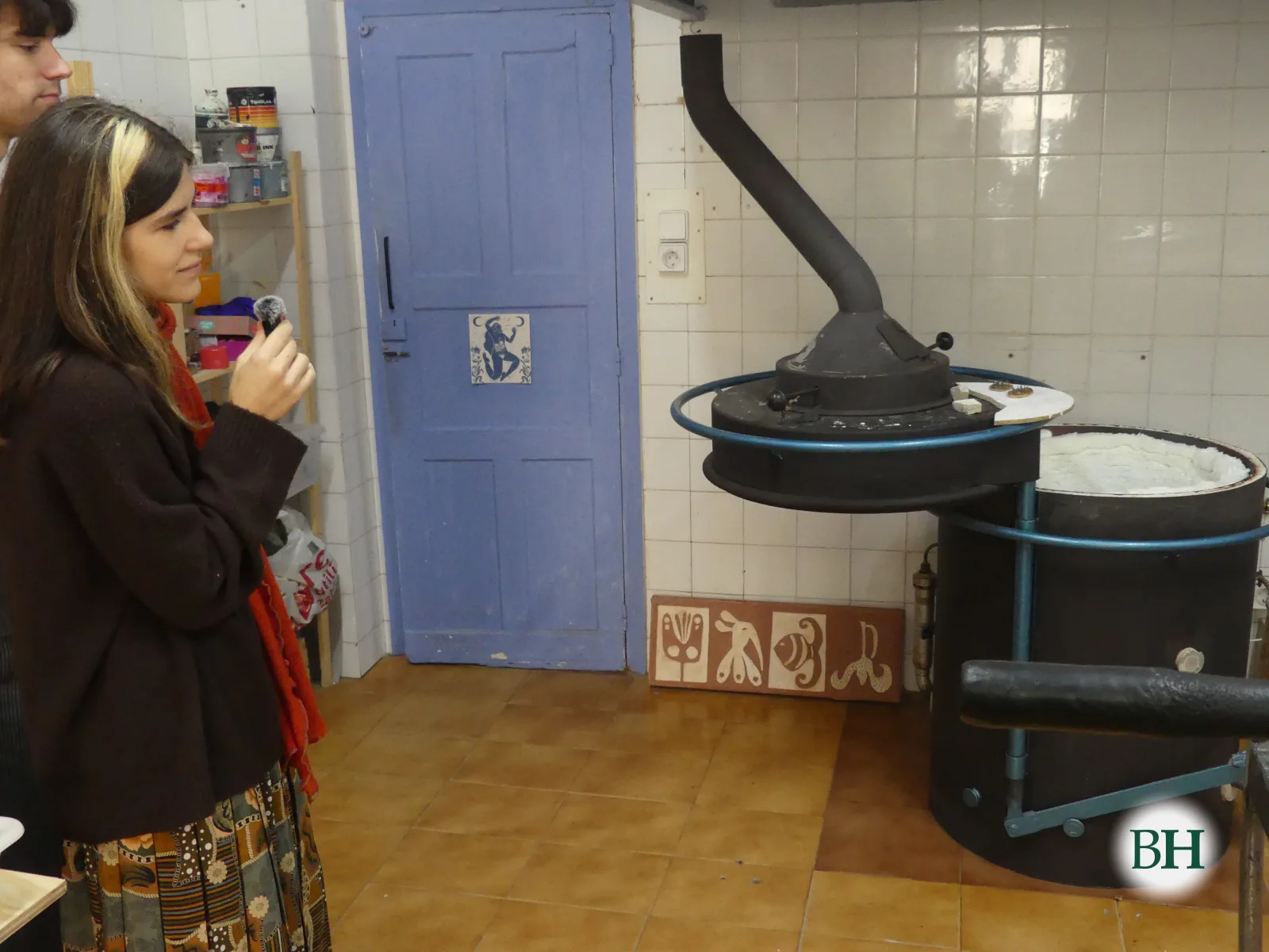 Andrea Aguafría, cun micrófono na man, diante dun forno de cerámica de gas, cilíndrico e negro, coa tapa aberta e unha cheminea que sobe cara á campá.