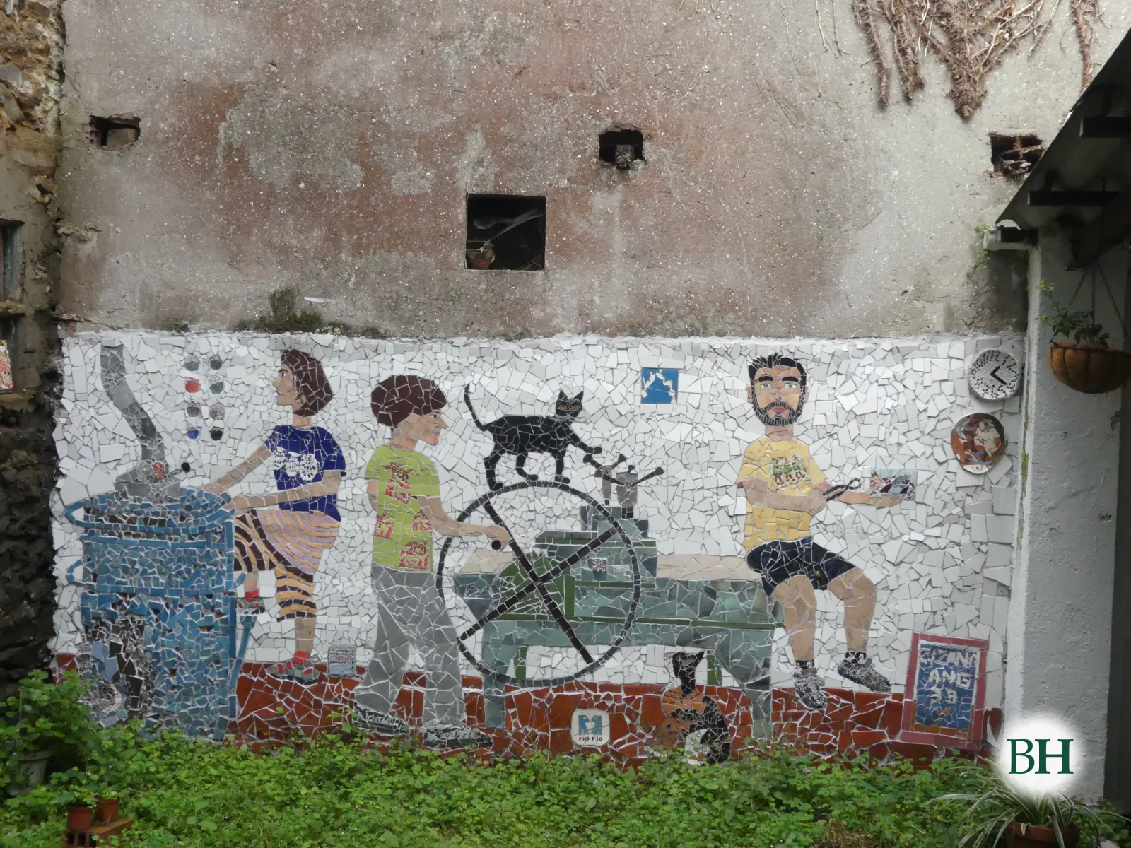 Mosaico na parede do patio do estudo, sobre a herba: unha muller xunto a un forno azul, un home que move a roda dun tórculo cun gato negro enriba, outro gato sentado debaixo e un home con barba que fai mosaico.