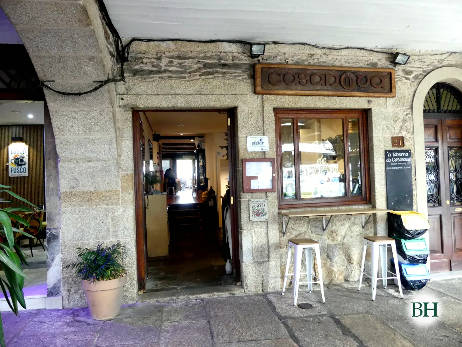 Entrada do Restaurante Casanova baixo os soportais de pedra, cun letreiro de madeira enriba da porta, unha xanela con mostrador e dous tallos brancos na beirarrúa.
