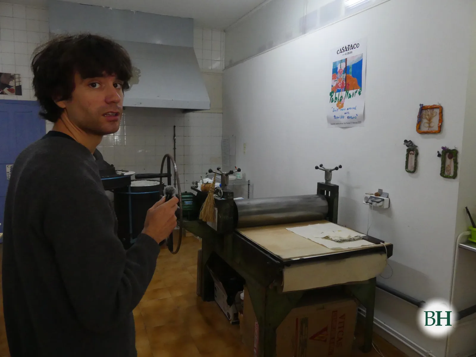 Pablo Naire, cun micrófono na man, ao lado dun tórculo verde co cilindro metálico e a pletina cuberta de papel. Na parede, o cartel dunha exposición súa en Casa Paco.