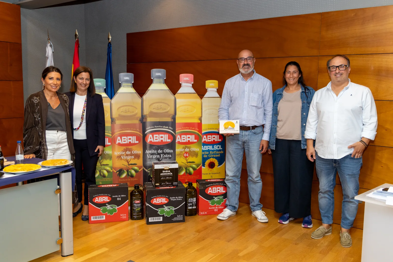 Seis persoas de pé xunto a botellas xigantes e caixas de Aceites Abril. Á esquerda, unha mesa con dúas tortillas en pratos.