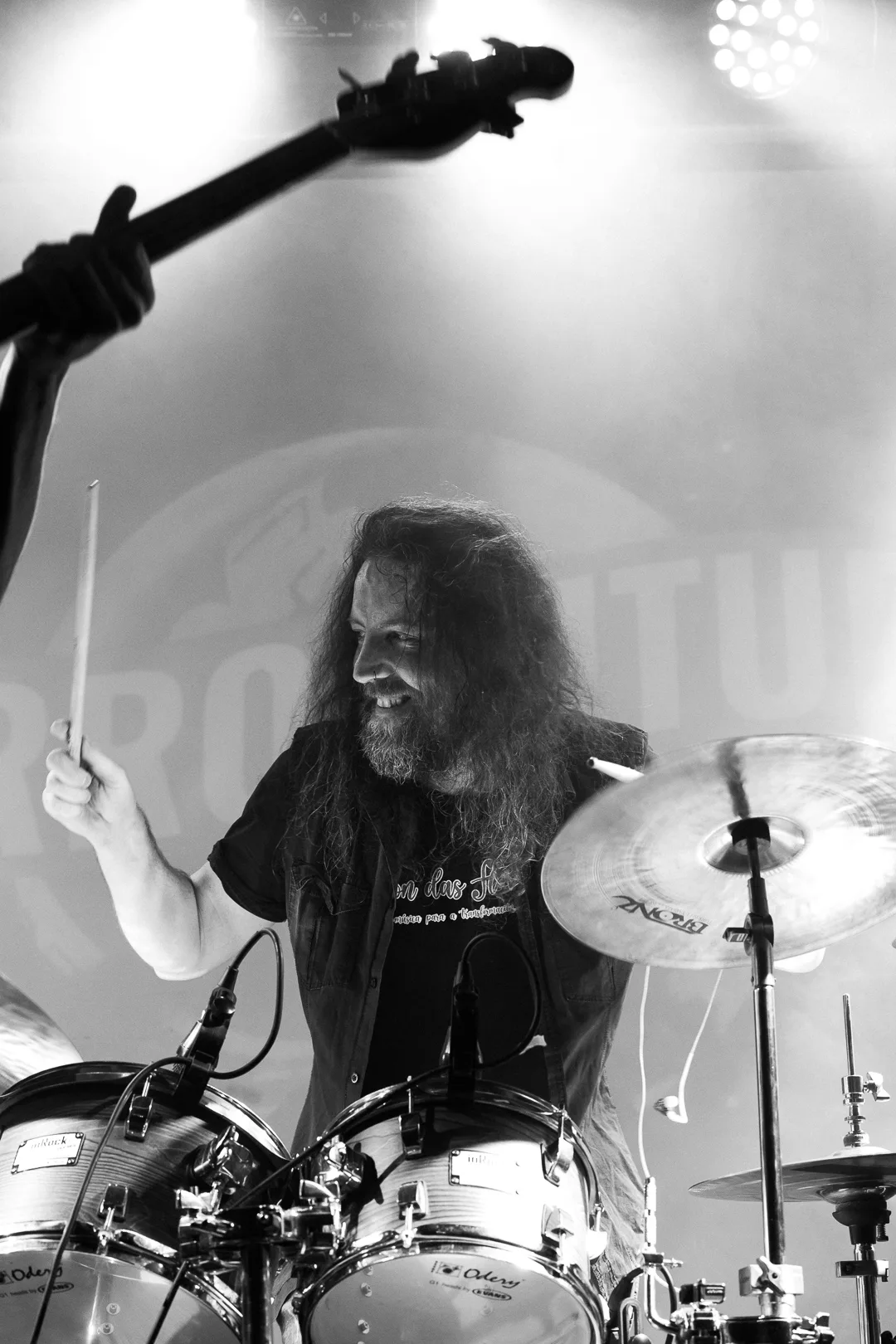 Suso Valcárcel, batería de Perro Futuro, sonríe mientras toca, con el mástil de un bajo en primer plano, en una foto de concierto en blanco y negro.
