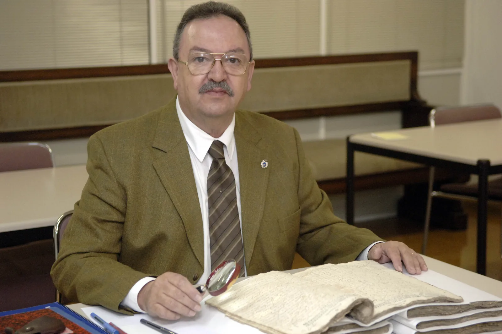 José Raimundo Núñez-Varela, con chaqueta e garavata, sentado diante duns legaxos antigos cunha lupa na man