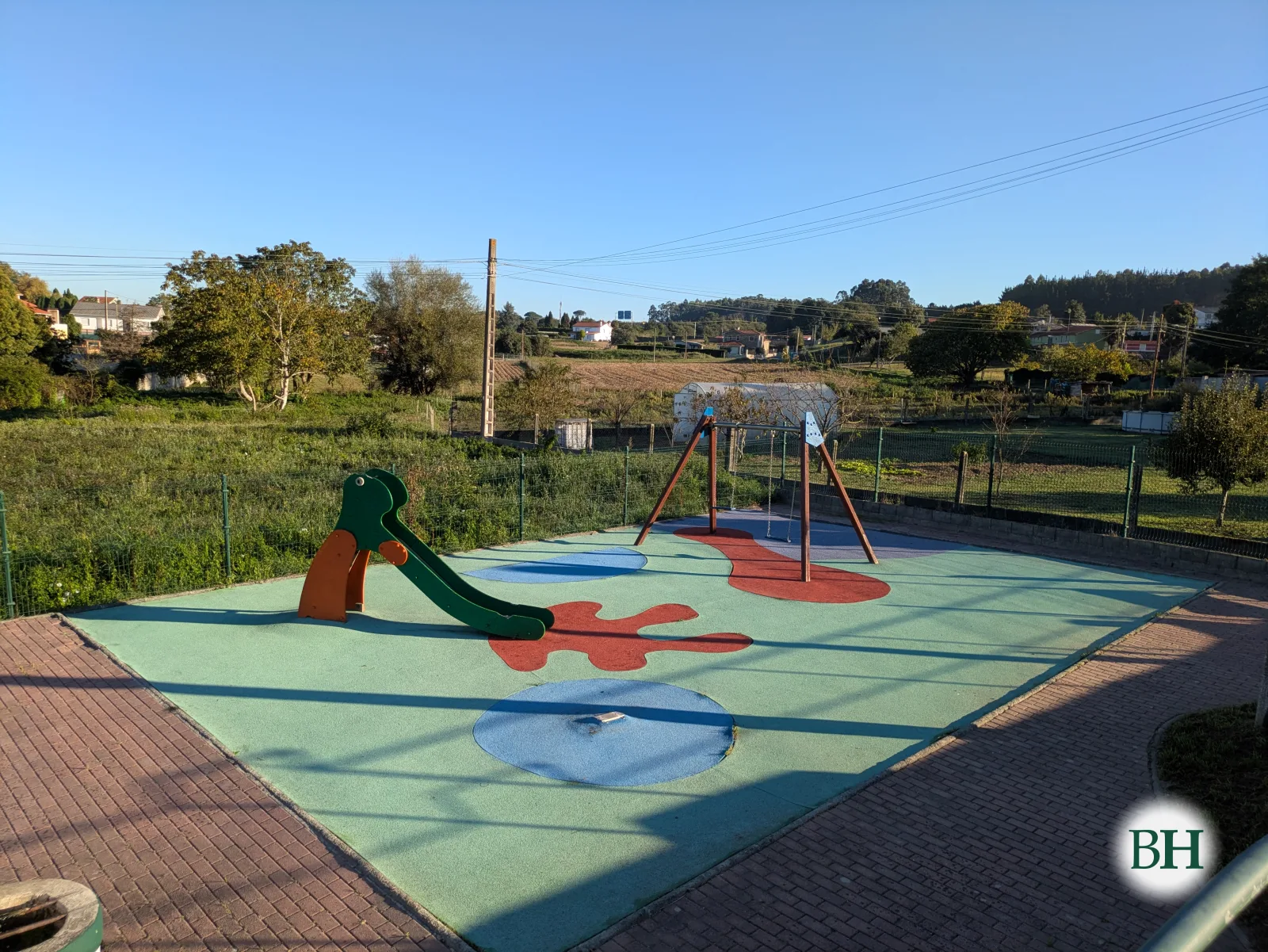 Parque infantil valado con tobogán e columpio sobre pavimento de caucho de cores, con leiras e vivendas ao fondo.