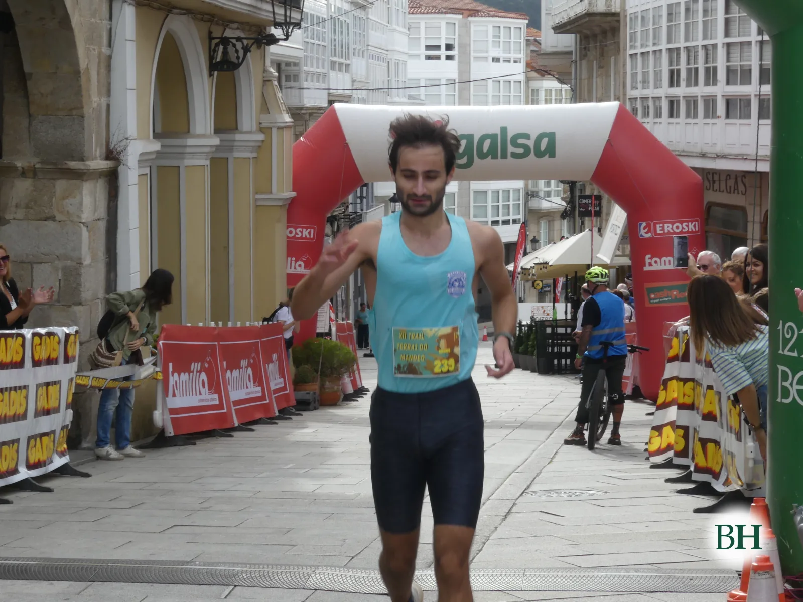 Corredor con camiseta azul de tirantes y el dorsal 239 llega a la meta saludando, con el arco hinchable al fondo.