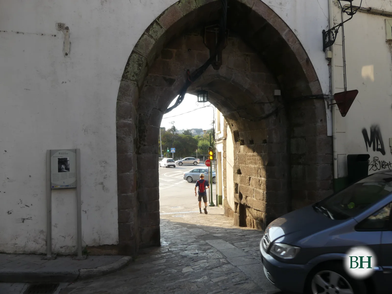 Vista máis próxima do mesmo arco de pedra, cun peón cunha mochila vermella ao fondo e un coche aparcado en primeiro plano.