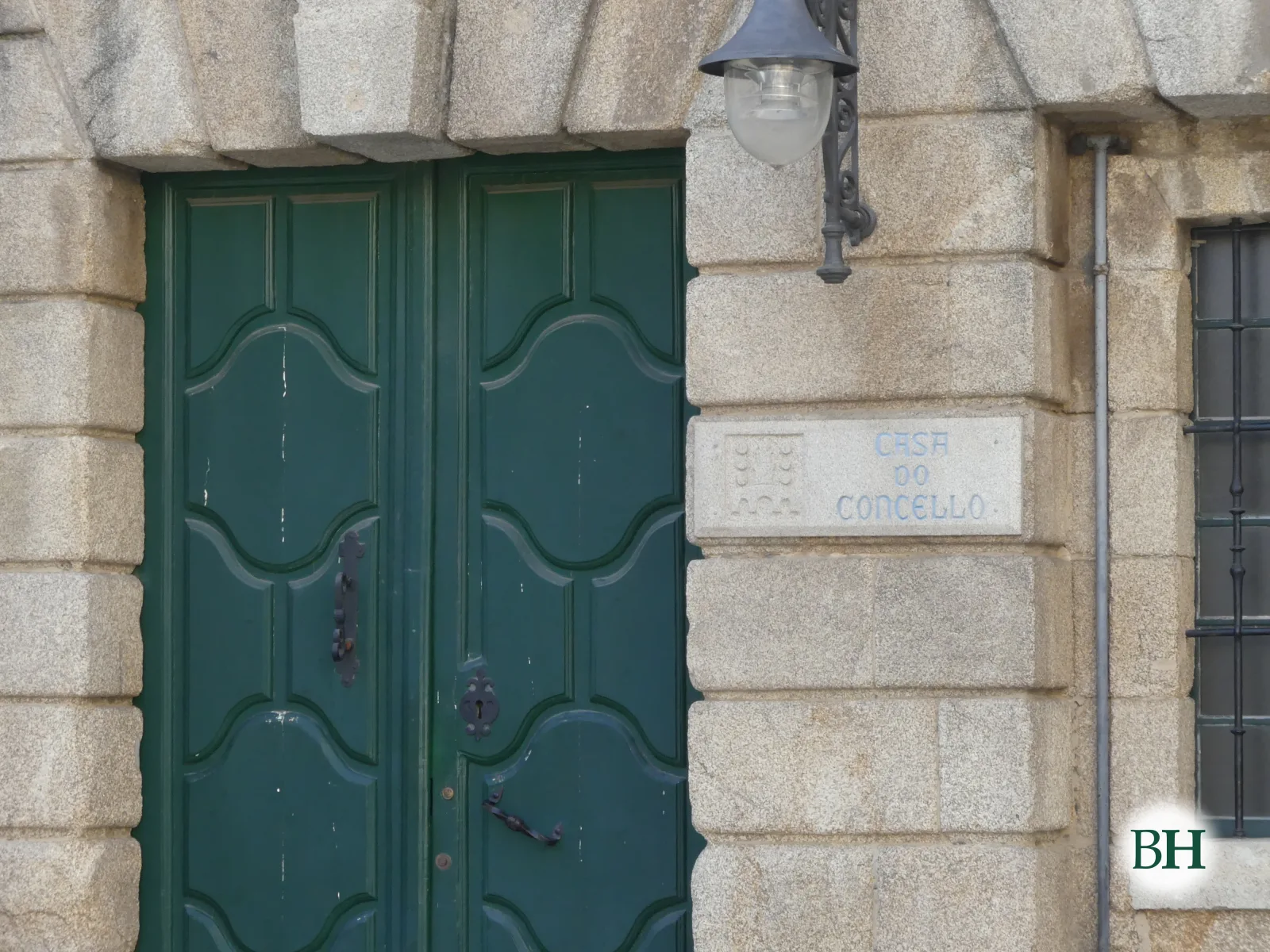 Puerta de doble hoja de madera verde con paneles curvos, enmarcada en sillares de granito, farol de hierro y placa con el texto CASA DO CONCELLO.