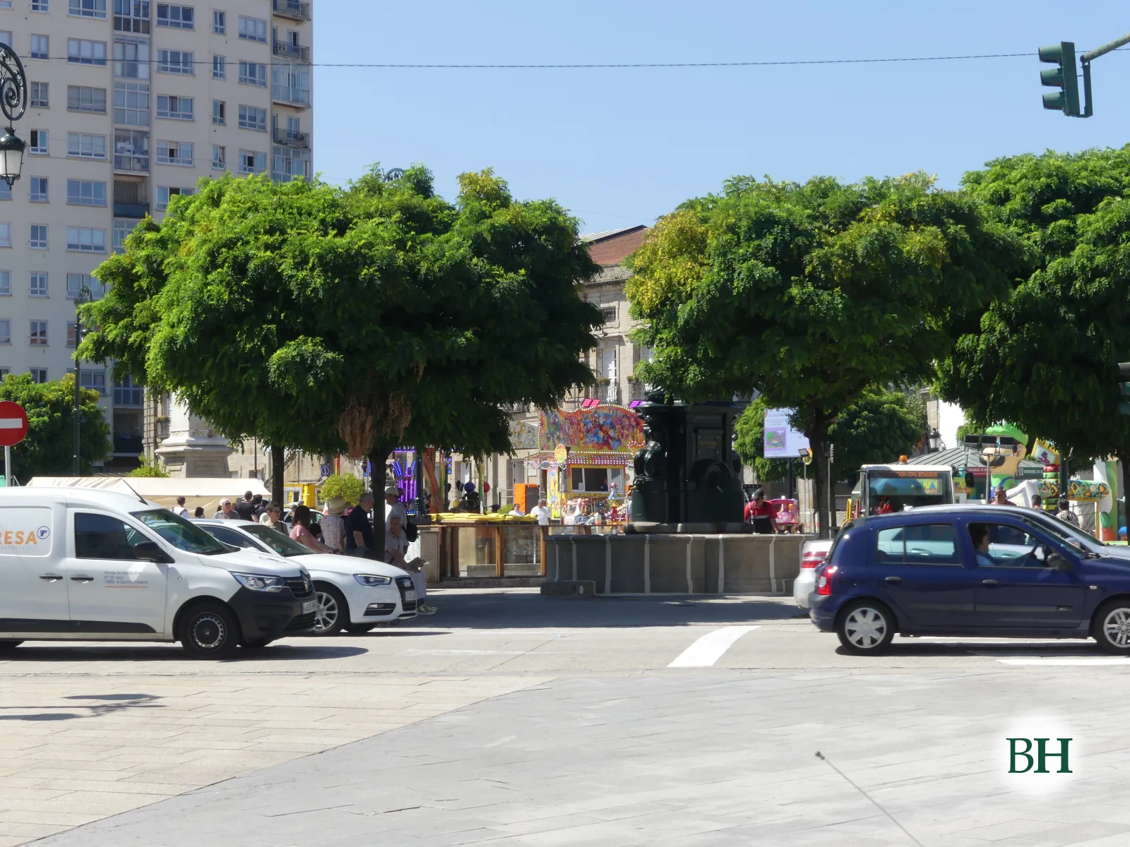 Plaza urbana con edificio de apartamentos al fondo, árboles frondosos, fuente central, puestos de feria y coches circulando bajo cielo despejado.