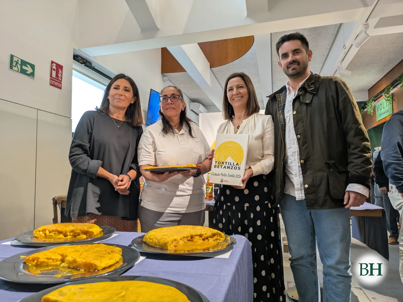 Sheila Delgado, do Mesón Café de Segundo, de polo branco e cun prato de tortilla, sorrinte con outras tres persoas detrás dunha mesa con mantel azul e cinco tortillas. Unha muller sostén un cadro amarelo de Gañador Mellor Tortilla 2026.