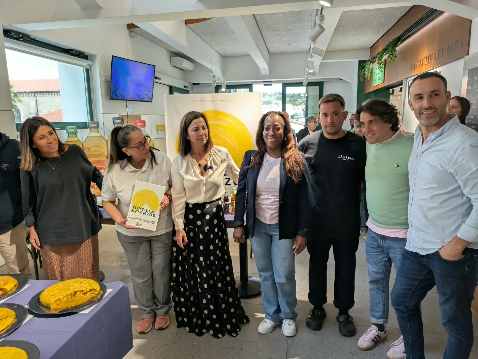 Nove persoas posan de pé e sorrintes nun local de paredes brancas e teito alto, diante dun cartel amarelo da Semana da Tortilla. Unha muller do centro sostén un diploma da Tortilla de Betanzos. Diante, unha mesa con mantel azul e cinco tortillas en
