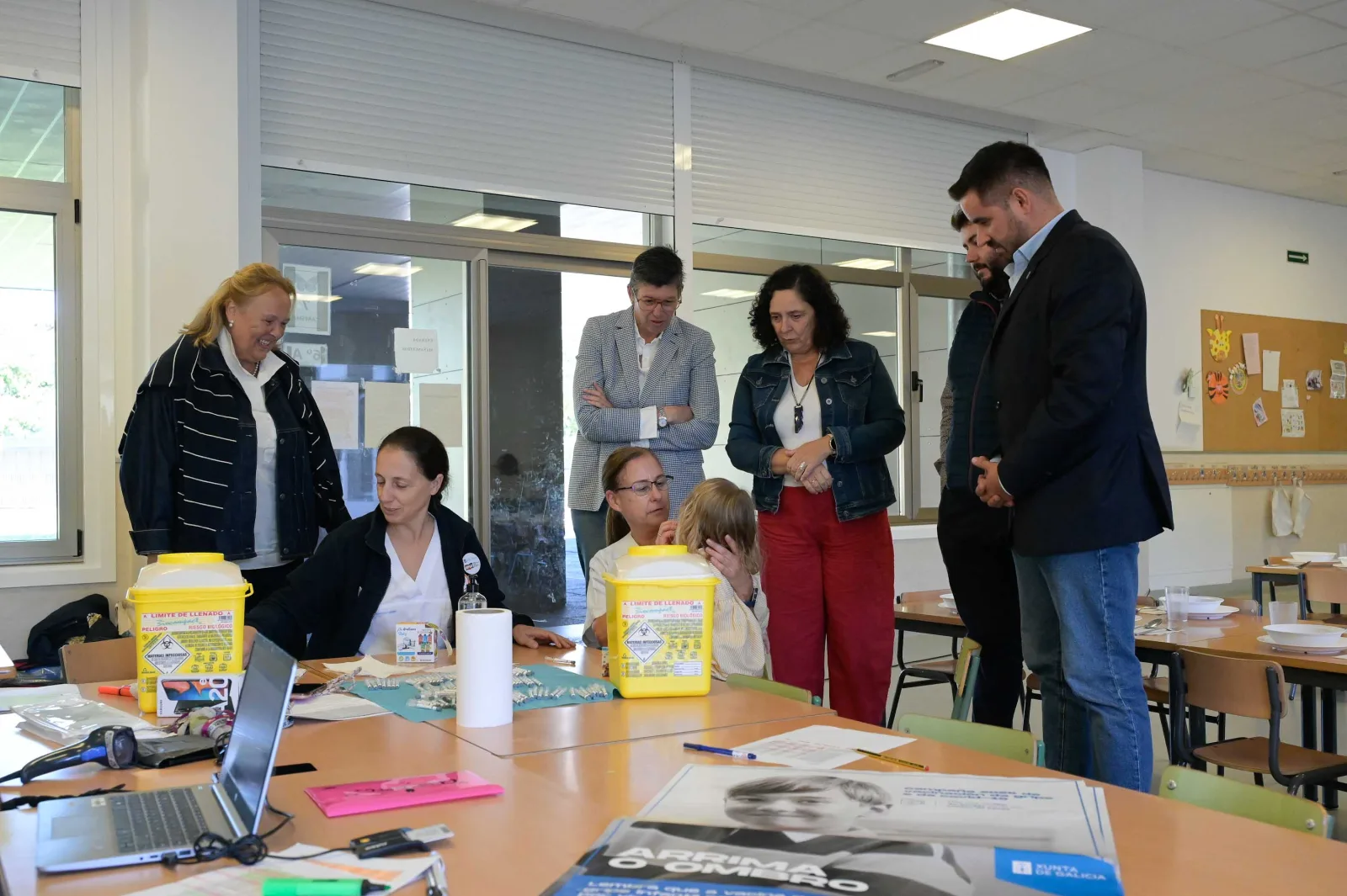 Unha enfermeira vacina contra a gripe a un dos pequenos no comedor do CEIP Vales Villamarín de Betanzos, xunto a outra sanitaria e diante das autoridades autonómicas que visitaron o colexio.
