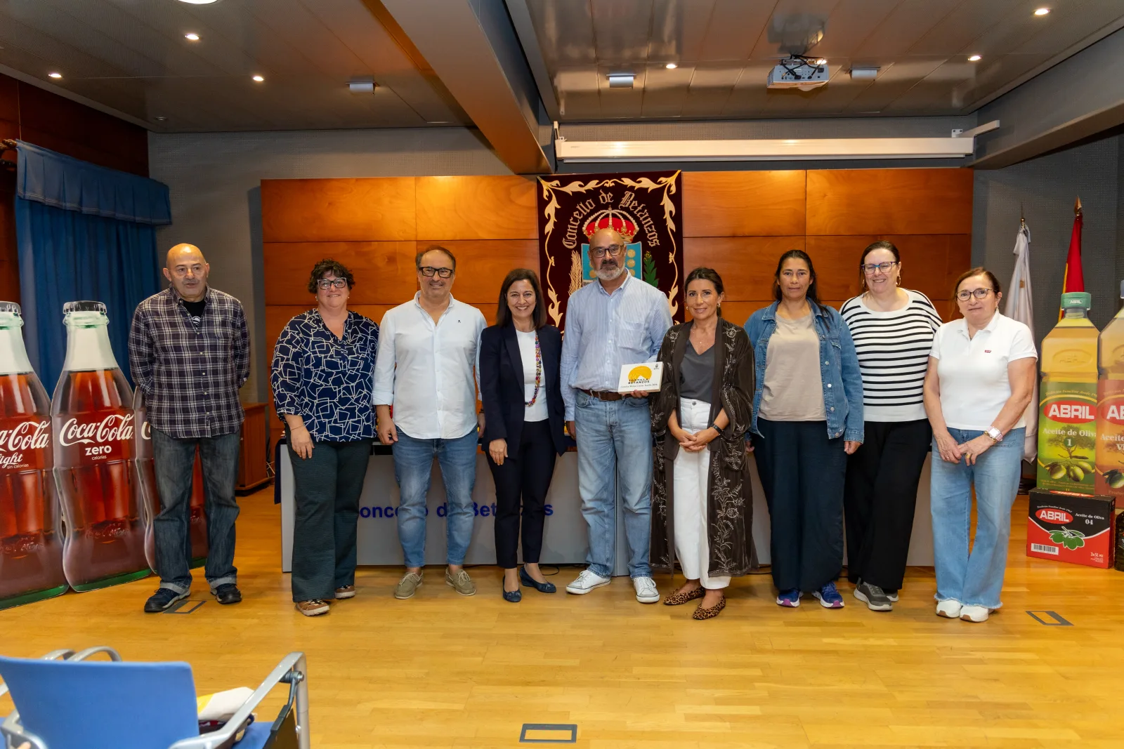 Foto de familia de once persoas de pé nun salón con parede de madeira, diante dun repostero do Concello de Betanzos. Un home do centro ten un diploma. Aos lados, botellas xigantes de refresco e de aceite.