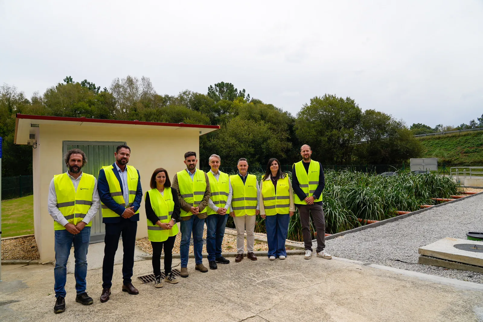 Oito persoas con chalecos reflectantes amarelos posan na inauguración do humidal de Viladesuso, diante dunha caseta e da balsa chea de espadana, na depuradora de Betanzos.